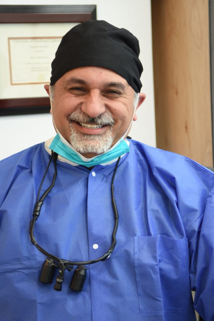 Dr. Omid Haroonian smiling in blue scrubs with a surgical mask lowered and loupes around his neck