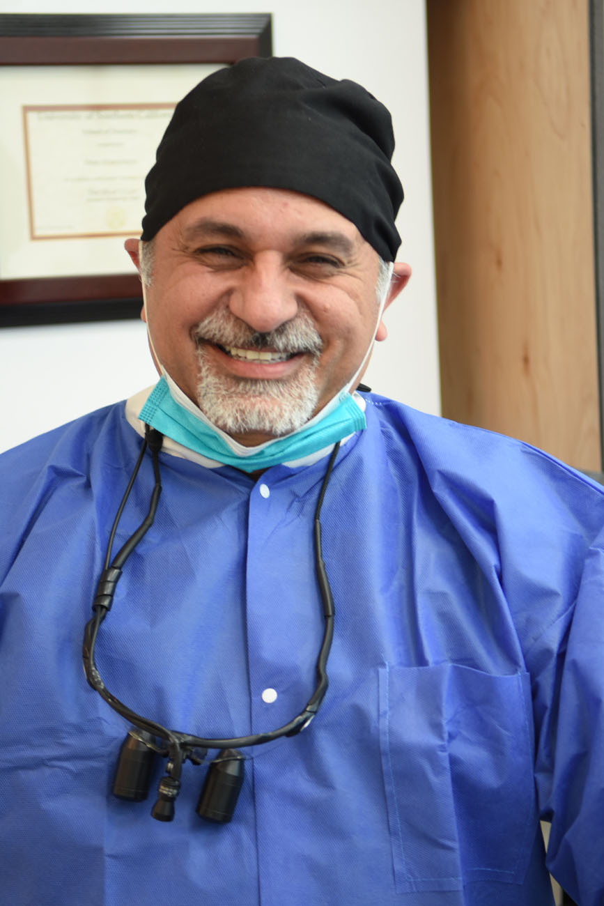 Dr. Omid Haroonian smiling in blue scrubs with a surgical mask lowered and loupes around his neck